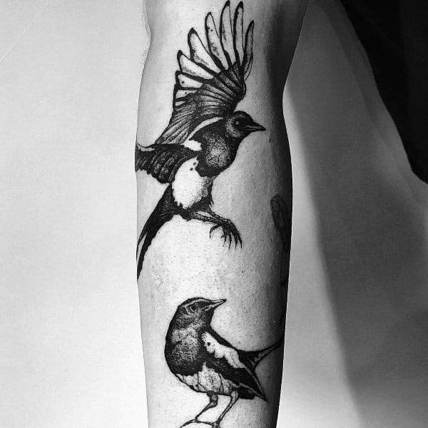 Black and gray magpie tattoo on forearm, featuring one bird in flight and another perched, creating a dynamic and elegant design.