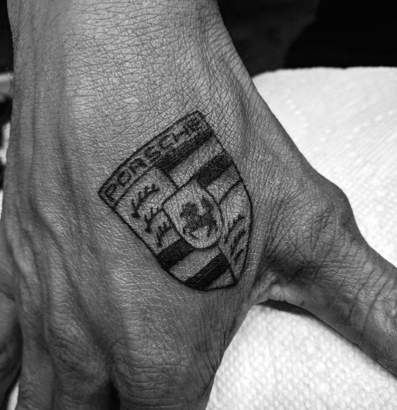 Black ink Porsche emblem tattoo on the back of a man's hand, symbolizing automotive enthusiasm.