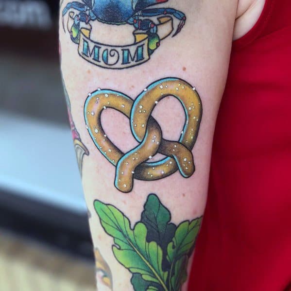 Golden pretzel tattoo with salt details and bold black outline on upper arm, surrounded by colorful traditional-style tattoos.