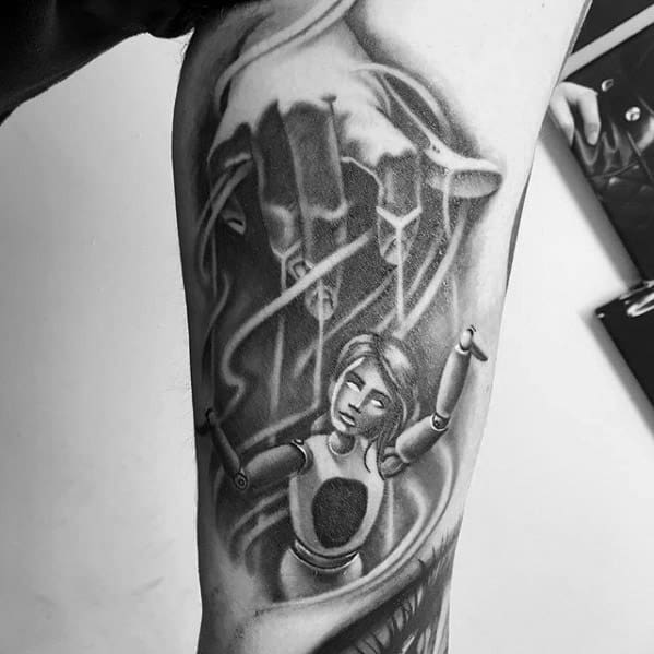 Black and grey puppet tattoo of a lifelike marionette controlled by a shadowy hand, symbolizing control and loss of autonomy.