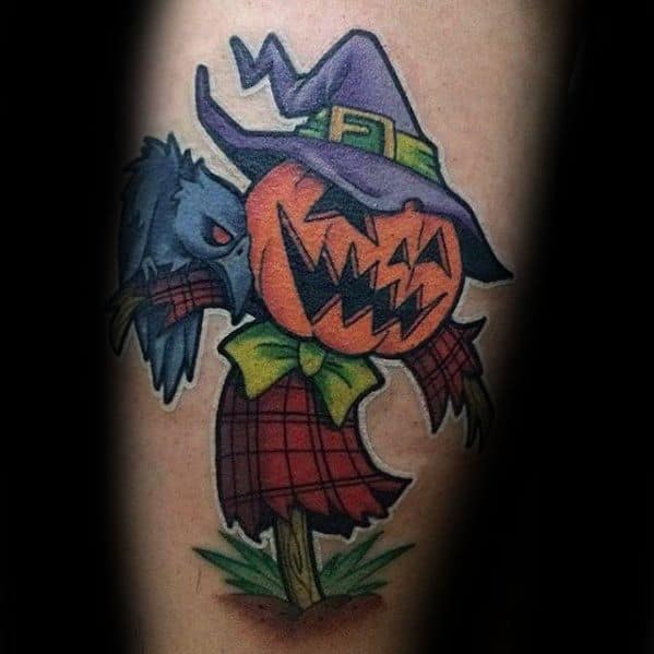 Colorful scarecrow tattoo with a grinning pumpkin head, purple witch hat, red plaid shirt, green bow, and cartoon-style crow.
