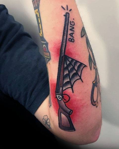 Minimalist shotgun tattoo on the forearm with a spiderweb detail and "BANG" text, featuring bold black outlines and a red background.