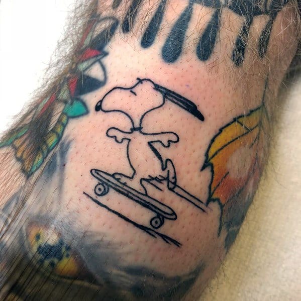 Black and white tattoo of Snoopy riding a skateboard with motion lines, placed on a person's leg among other colorful tattoos.