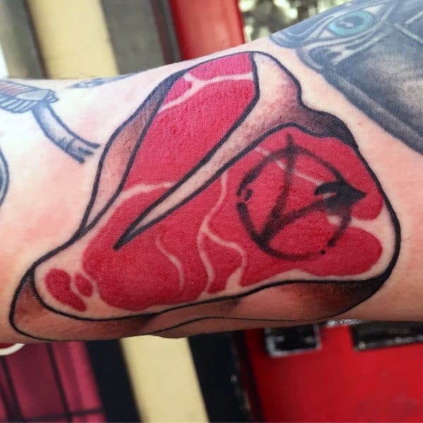 Tattoo of a detailed steak with marbling, featuring a bold anarchist symbol burned into the meat, creating a rebellious and unique design