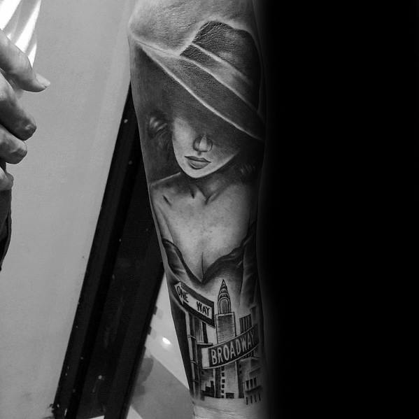 Black and grey tattoo of a mysterious woman in a wide-brimmed hat, with detailed Broadway street signs and city buildings below.