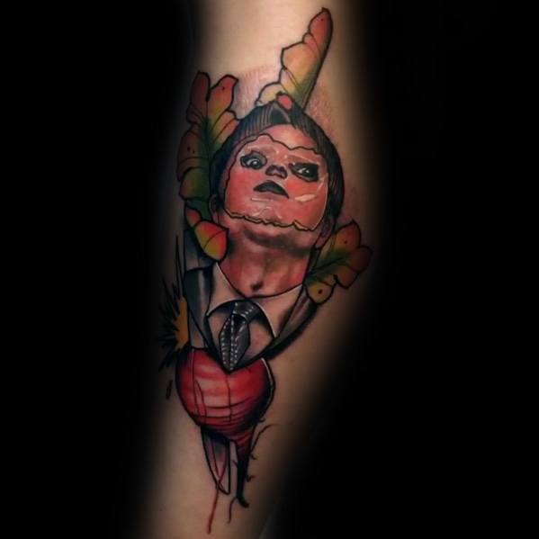 Tattoo of Dwight Schrute with a puppet-like face merging into a beet, surrounded by colorful leaves, blending realism and surrealism on an arm.