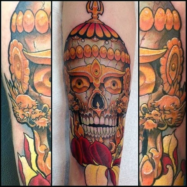 Bright Tibetan skull tattoo with golden-orange tones, crown-like headpiece, and floral elements wrapping around the lower arm.