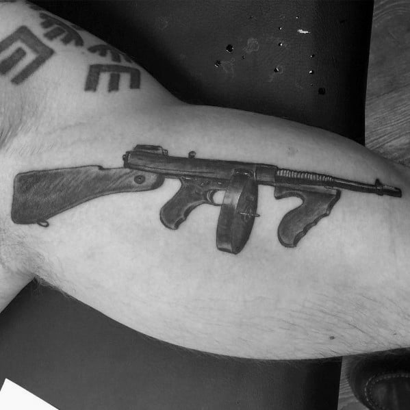 Realistic black and grey Tommy gun tattoo on the inner bicep, featuring fine shading and detailed design for a classic firearm look.