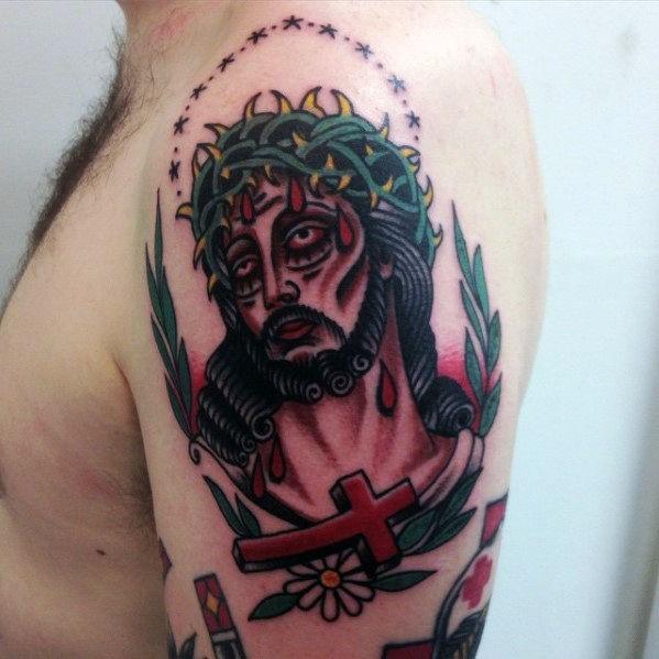 Traditional-style upper arm tattoo of Jesus with a crown of thorns, surrounded by leaves, stars, a red cross, and a white flower.