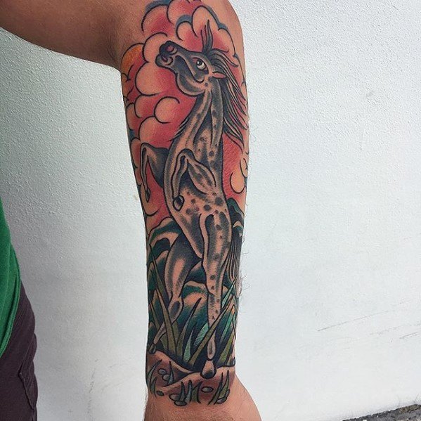 Traditional tattoo of a spotted rearing horse surrounded by grass and pink-orange clouds, stretching along the forearm.