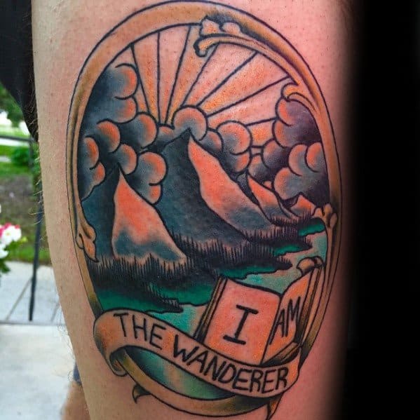 Framed mountain and river tattoo with dramatic clouds, rising sun, and an open book reading "I Am the Wanderer."