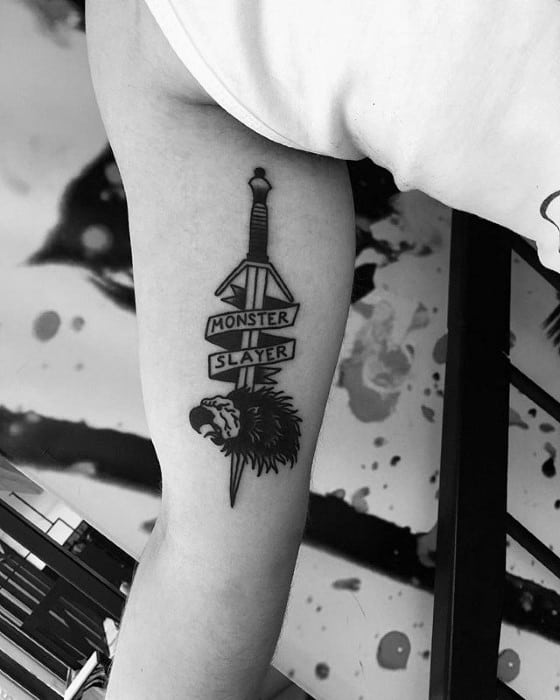 Minimalist Witcher tattoo on the upper arm featuring a sword, wolf medallion, and bold text reading “Monster Slayer.”