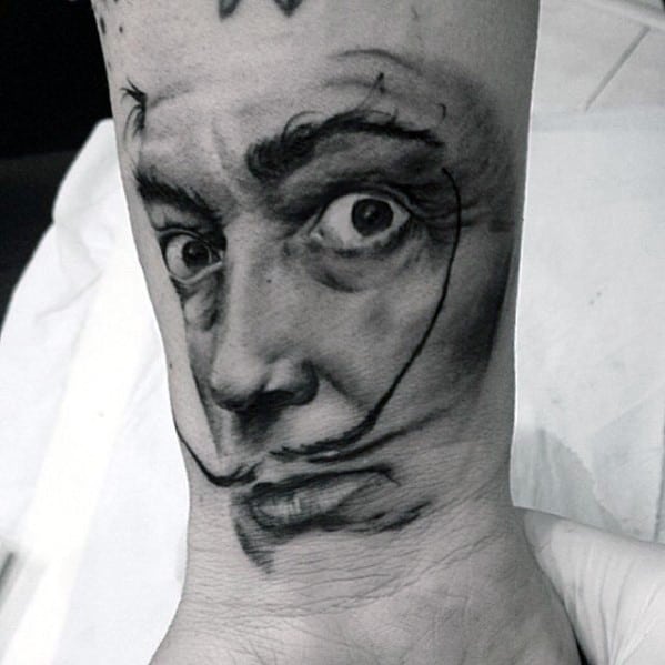Black and grey wrist tattoo of Salvador Dali featuring his wide-eyed stare and signature mustache in a dramatic, sketch-style portrait.