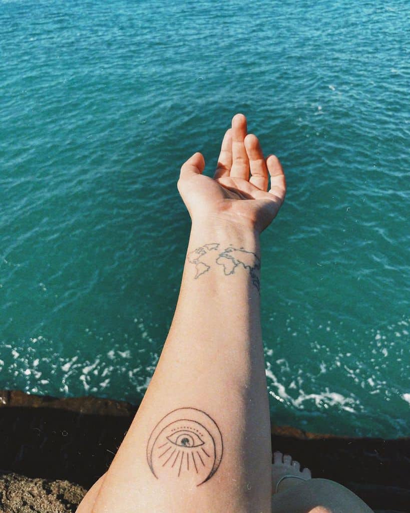 Forearm tattoo of an all-seeing eye within a semi-circle, radiating lines, and a minimalist world map, with an ocean backdrop.