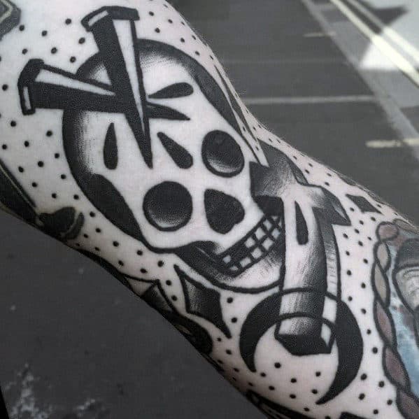 Black and white tattoo of a skull with nails in the head and a cross through the eye, surrounded by bold lines and dot shading.