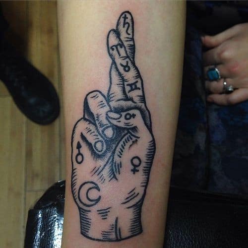 Bold blackwork tattoo of a crossed-finger hand adorned with astrological and alchemical symbols, placed on the forearm.