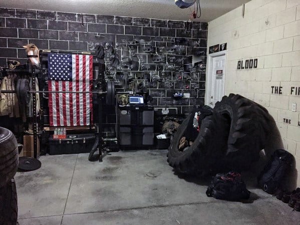 Rustic garage gym with large tires, barbell setup, chalkboard wall, and American flag decor.