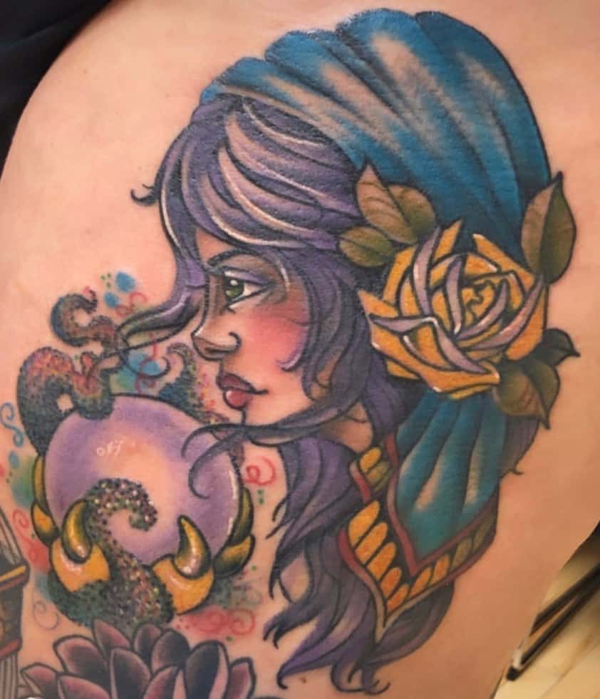 Colorful tattoo of a gypsy woman with a blue headscarf, golden rose, and a mystical crystal ball with swirling details on the thigh.