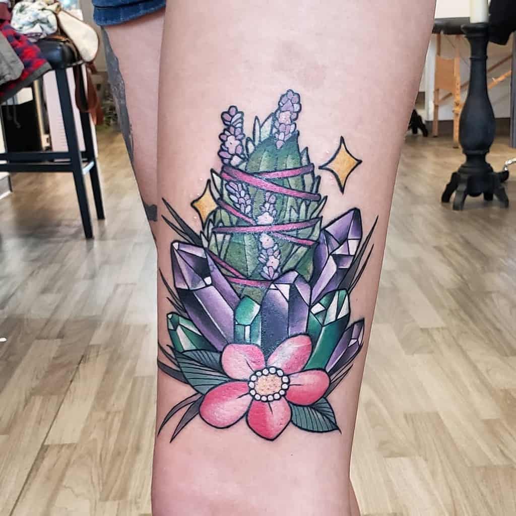 A colorful tattoo of lavender wrapped in pink ribbons, surrounded by geometric crystals and a pink flower, with sparkling accents.