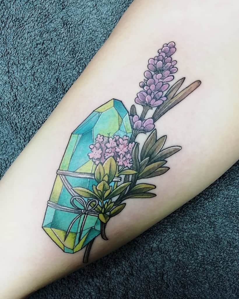 A colorful tattoo of a geometric crystal wrapped with a ribbon, surrounded by lavender and greenery, placed on the inner arm.