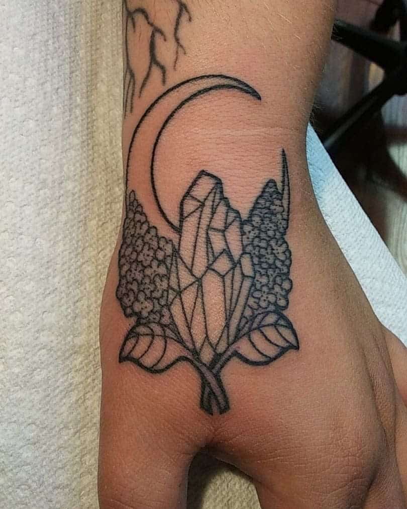 Black ink tattoo on the hand featuring a lilac branch with a crystal cluster and a crescent moon, blending botanical and mystical elements.