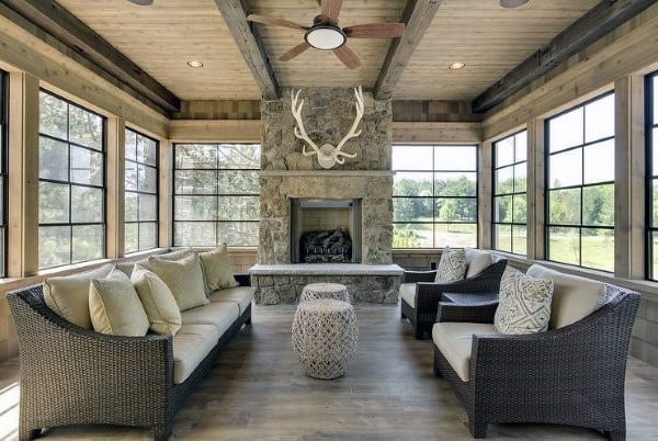Bright sunroom with stone fireplace, antler decor, wicker furniture, and wooden ceiling beams.