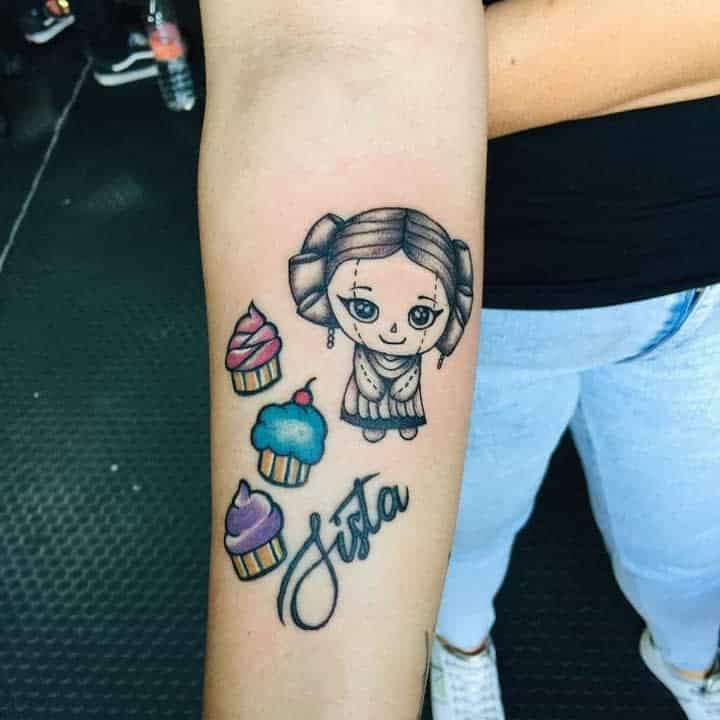 Colorful tattoo featuring a cute chibi-style doll girl, three cupcakes, and the word 'Sista' in cursive