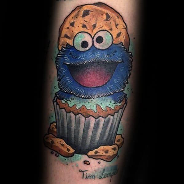 Playful Cookie Monster tattoo with a wide grin, sitting inside a cupcake topped with a giant cookie and surrounded by crumbs.