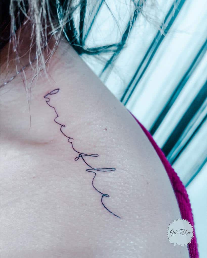 Elegant script tattoo spelling "breathe" on a person's shoulder, with strands of hair partially visible