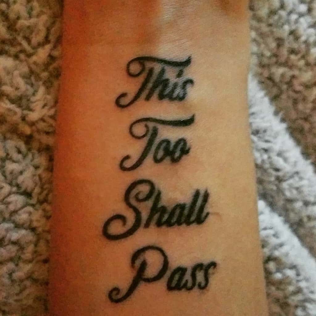 A tattoo on the forearm that displays the phrase "This Too Shall Pass" in a bold, elegant font with stacked lettering.
