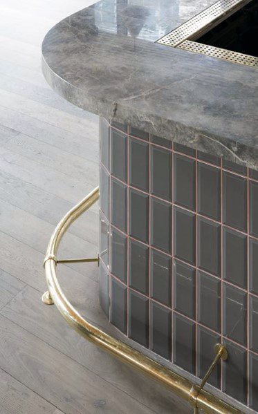 Curved brass foot rail attached to a grey tiled bar with a marble countertop and light wood flooring.