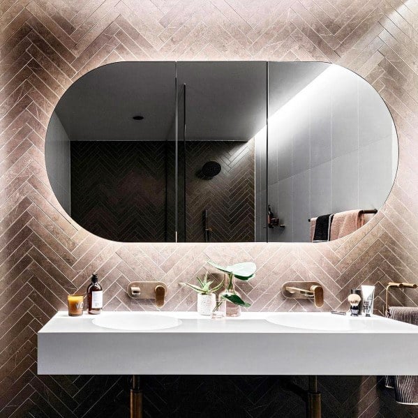 Oval LED backlit mirror with double sinks and gold faucets in modern bathroom design.