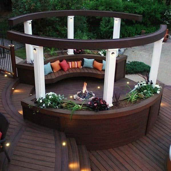 Outdoor circular deck with a fire pit, bench seating, colorful cushions, and surrounding plants