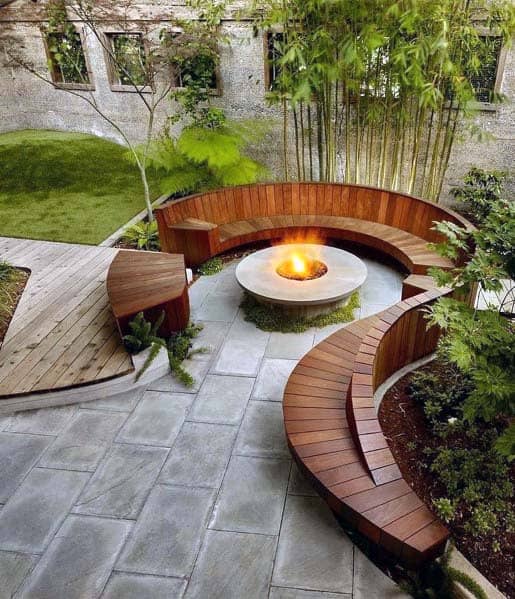 Circular wooden benches curve around a central fire pit in a modern outdoor garden space with trees and grass