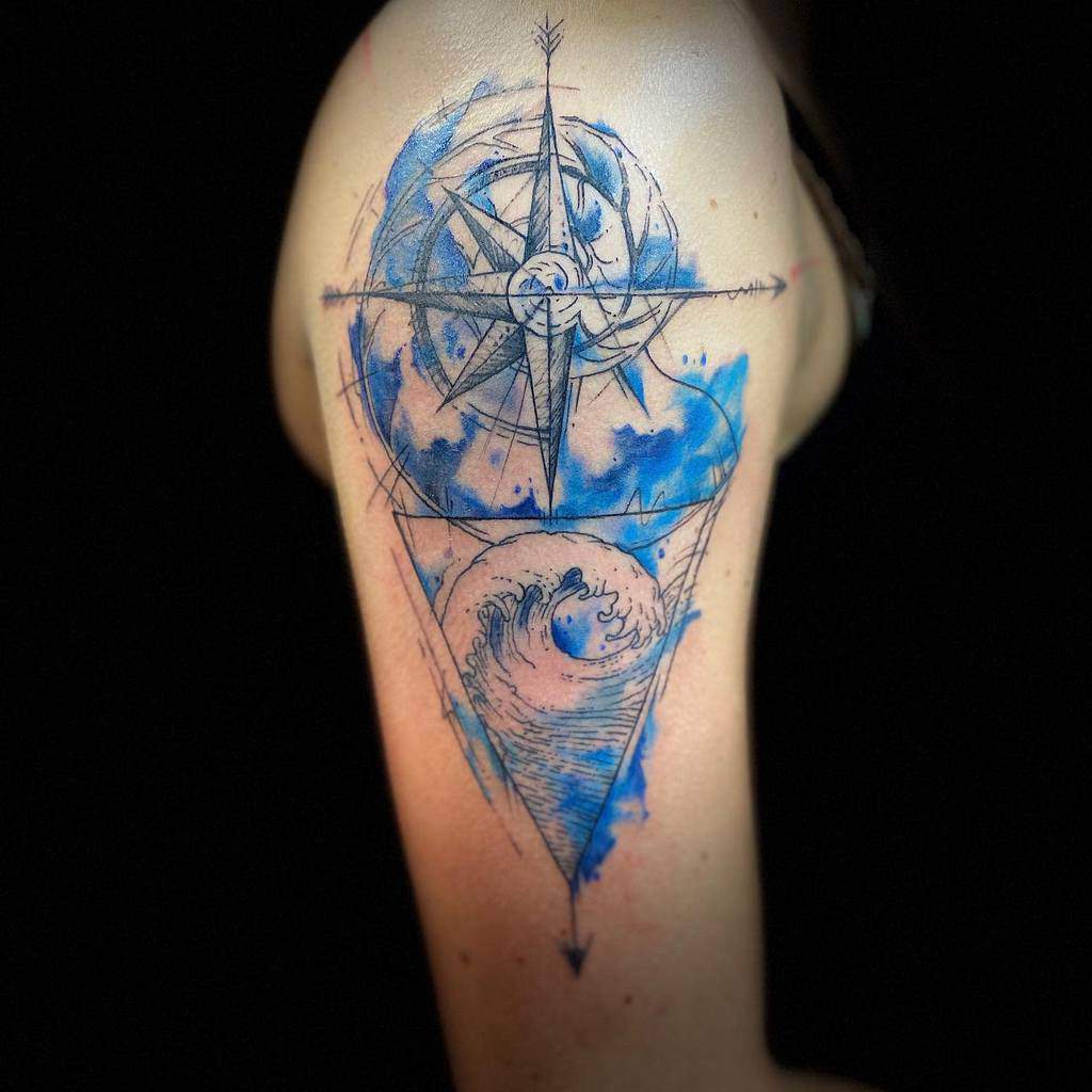 Watercolor-style tattoo featuring a detailed compass rose above a crashing ocean wave, enclosed in a geometric frame with blue ink splashes