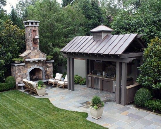 Inviting pavilion with a metal roof, outdoor kitchen, and stone fireplace, surrounded by lush greenery.