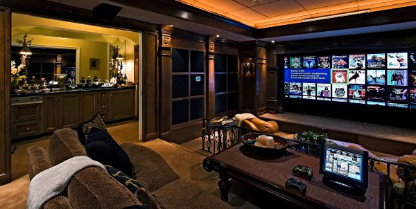 Cozy home theater features large screen, brown sofas, gaming controllers, and open kitchen backdrop