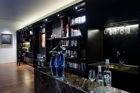 Modern home bar with dark wood bar shelves, a ladder, wine glasses, and drinks on a marble counter; painting on the wall
