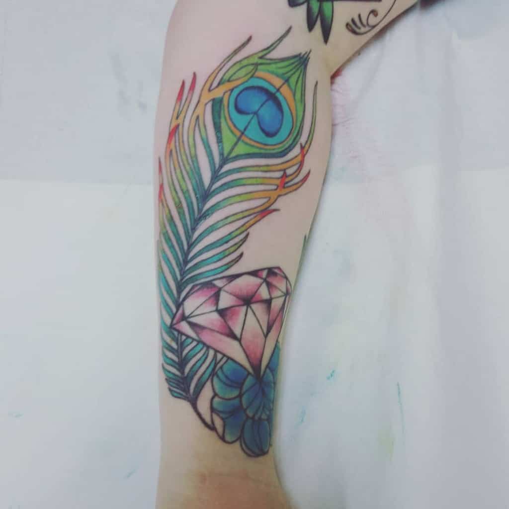Colorful tattoo on the forearm featuring a vibrant peacock feather, a pink gemstone, and blue flowers with fine linework and shading.