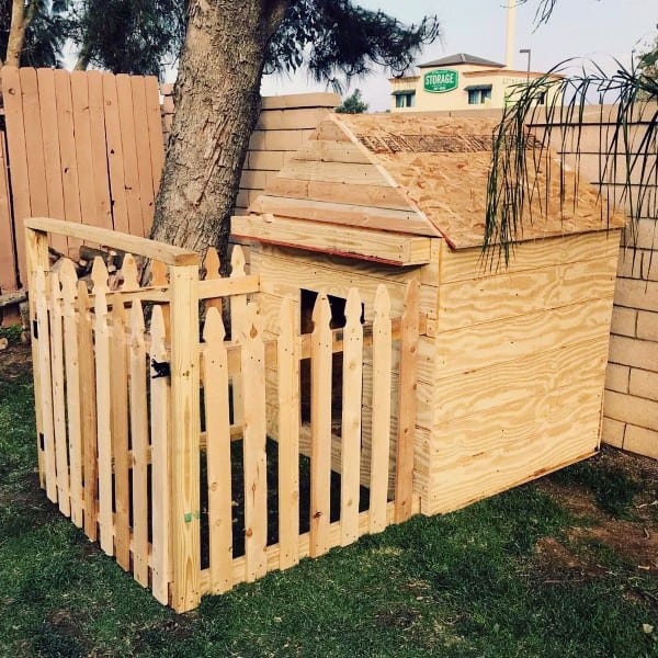 Custom wooden dog house with a sloped roof, enclosed by a picket fence, set in a grassy yard near a tree and a wall