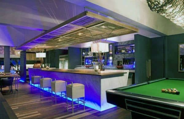 Modern bar interior with illuminated counter, white chairs, a pool table, and ambient blue bar lighting