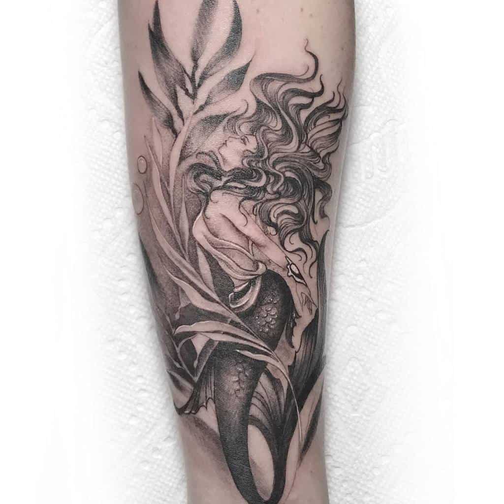 Black and grey tattoo of a mermaid with flowing hair, surrounded by seaweed, with intricate shading and fine details on the forearm.