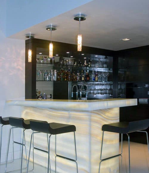 Modern bar with illuminated counter, three barstools, an array of bottles, glassware, and hanging lights