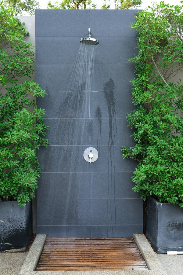 Modern outdoor shower with sleek dark tiles, wooden floor mat, and lush green potted plants.