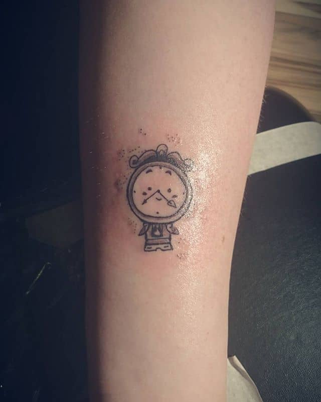 Minimalist black ink tattoo of Cogsworth from Beauty and the Beast, drawn in a simple, cartoonish style on the forearm.