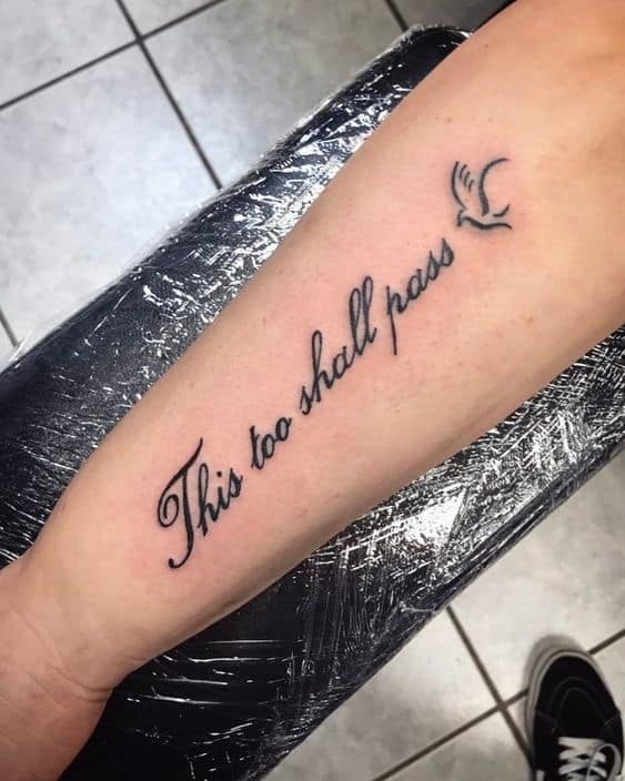 A "This Too Shall Pass" tattoo in cursive with a flying bird design, on a person's forearm.