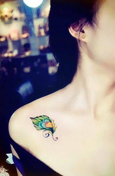 Small and colorful peacock feather tattoo on the collarbone, featuring vibrant green, blue, and yellow hues with elegant swirl details.
