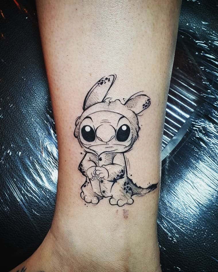 Black and white tattoo of Stitch from Lilo & Stitch wearing a bunny onesie, sitting on the forearm with a sad expression.