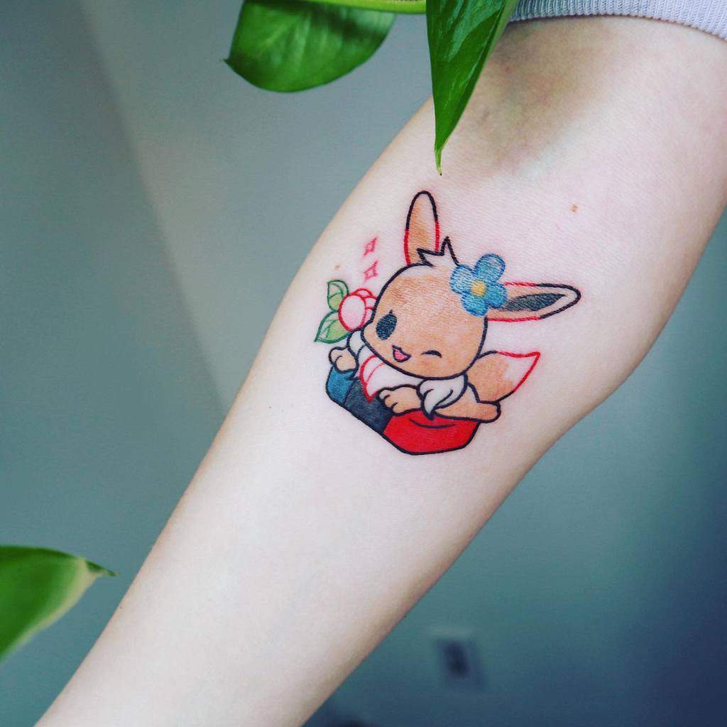 Charming Eevee tattoo with a blue flower and a joyful pose, sitting in a box with soft pastel colors and sparkles