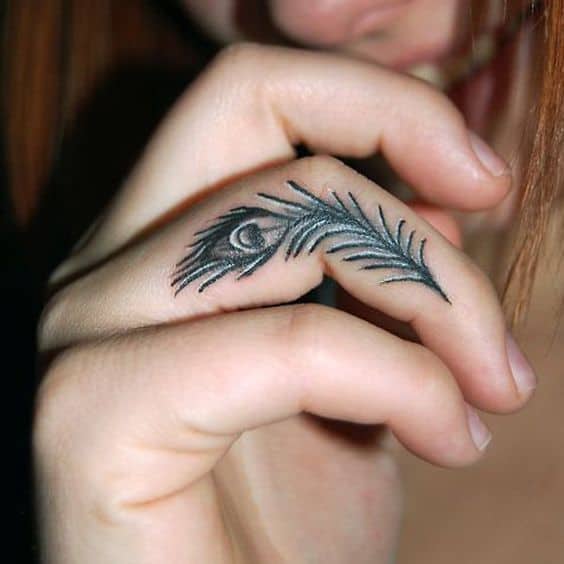 Small black ink peacock feather tattoo on the side of the finger, featuring fine linework and subtle shading for an elegant minimalist look.